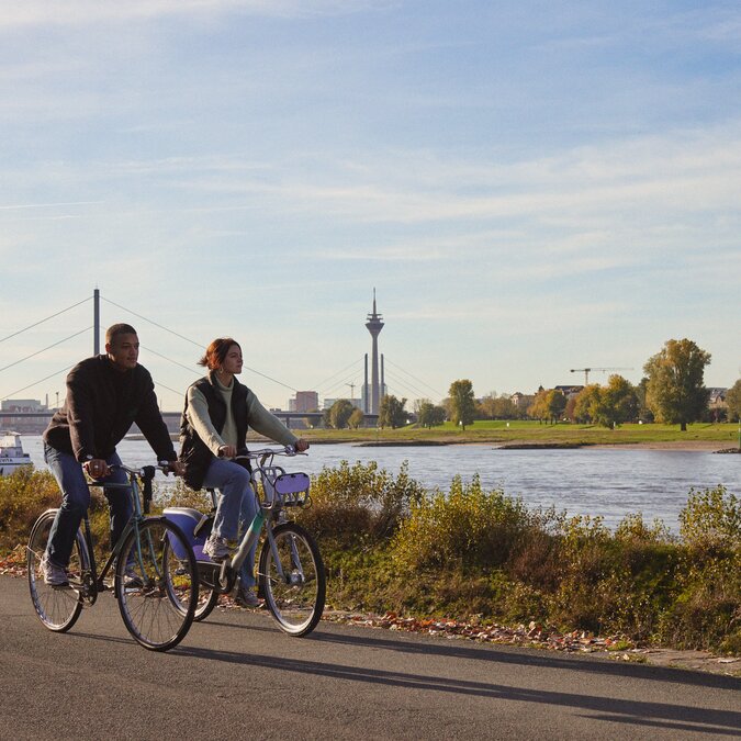 Zwei Personen radeln am Rheinufer mit Blick auf den Rheinturm. | © Düsseldorf Tourismus – React-EU