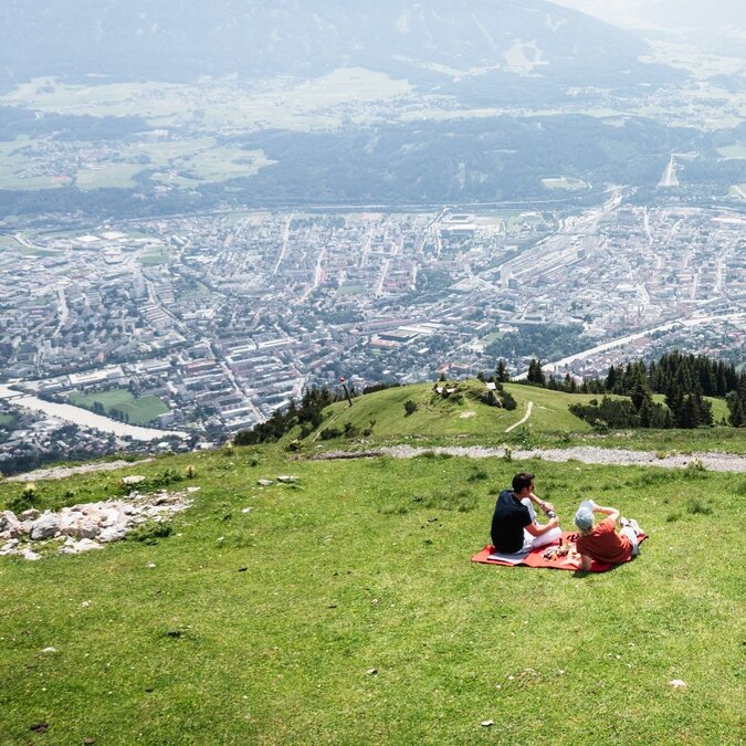 Zwei Personen sitzen auf einer Decke auf einer grünen Bergwiese oberhalb von Innsbruck. Unterhalb liegt die Stadt mit ihren Gebäuden und Strassenzügen, dahinter erstrecken sich das Inntal und umliegende Bergketten. | © Innsbrucker Nordkettenbahnen, L-Jaenichen