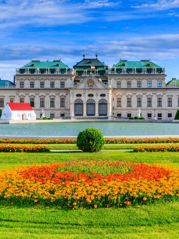 Barockes Schloss Belvedere in Wien mit grünen Kupferdächern, davor ein Wasserbecken und symmetrisch angelegte Blumenbeete in einem gepflegten Schlossgarten unter blauem Himmel. | © shutterstock 678034990