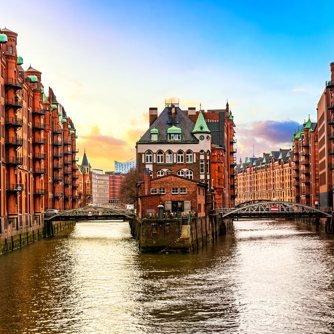 Das historische Wasserschloss in der Hamburger Speicherstadt steht zwischen zwei Kanälen und ist von roten Backsteinspeichern umgeben. Brücken überspannen das Wasser, während die Elbphilharmonie im Hintergrund zu erkennen ist. | © shutterstock_1946862949